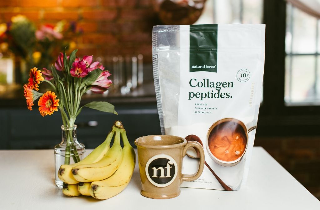 a handmade natural force mug containing coffee with collagen peptides on a counter beside a bag of natural force collagen peptides and bananas