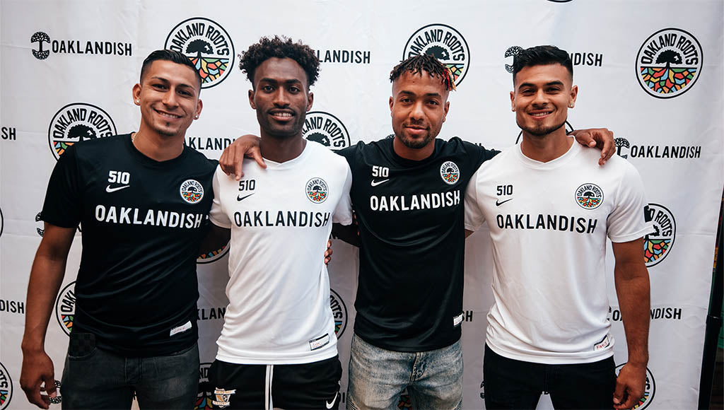 Four guys in front of step and repeat wearing Roots soccer jerseys