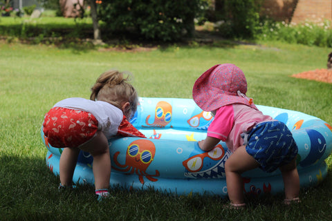 Two girls leaning into a blow up pool in the grass wearing Beau and Belle Littles swim diapers in the flamingo and green arrow prints. 