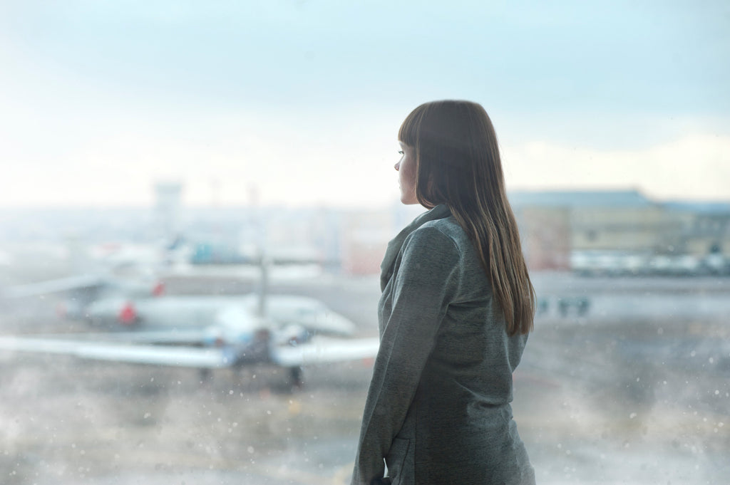 Woman in a green jackert standing in front of an airport window looking out at the airplanes on the tarmac